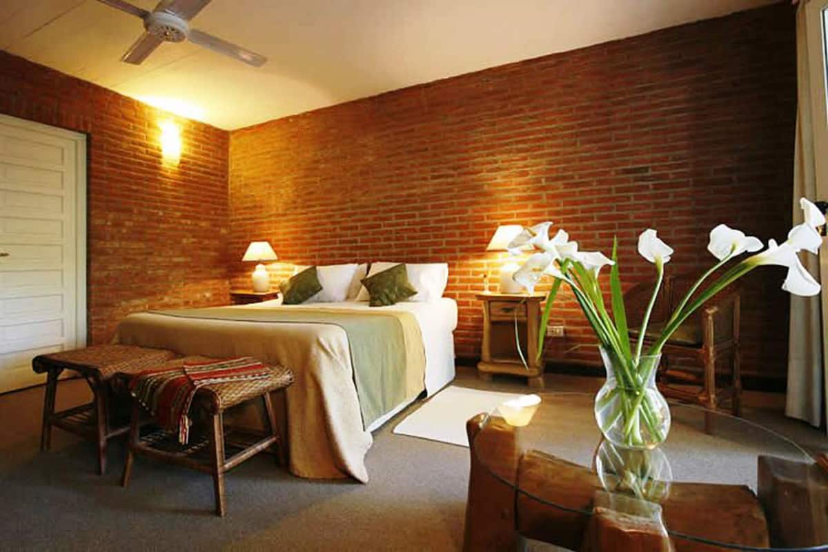 Quarto com parede de tijolos, cama, cadeira, mesa e um vaso com flores.