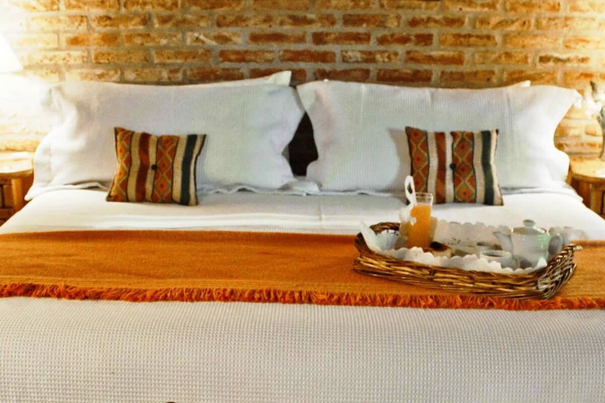 Cama com lençóis brancos, duas almofadas com desenhos estampados, um cesto de vime com um copo de suco de laranja e uma parede de tijolos ao fundo.