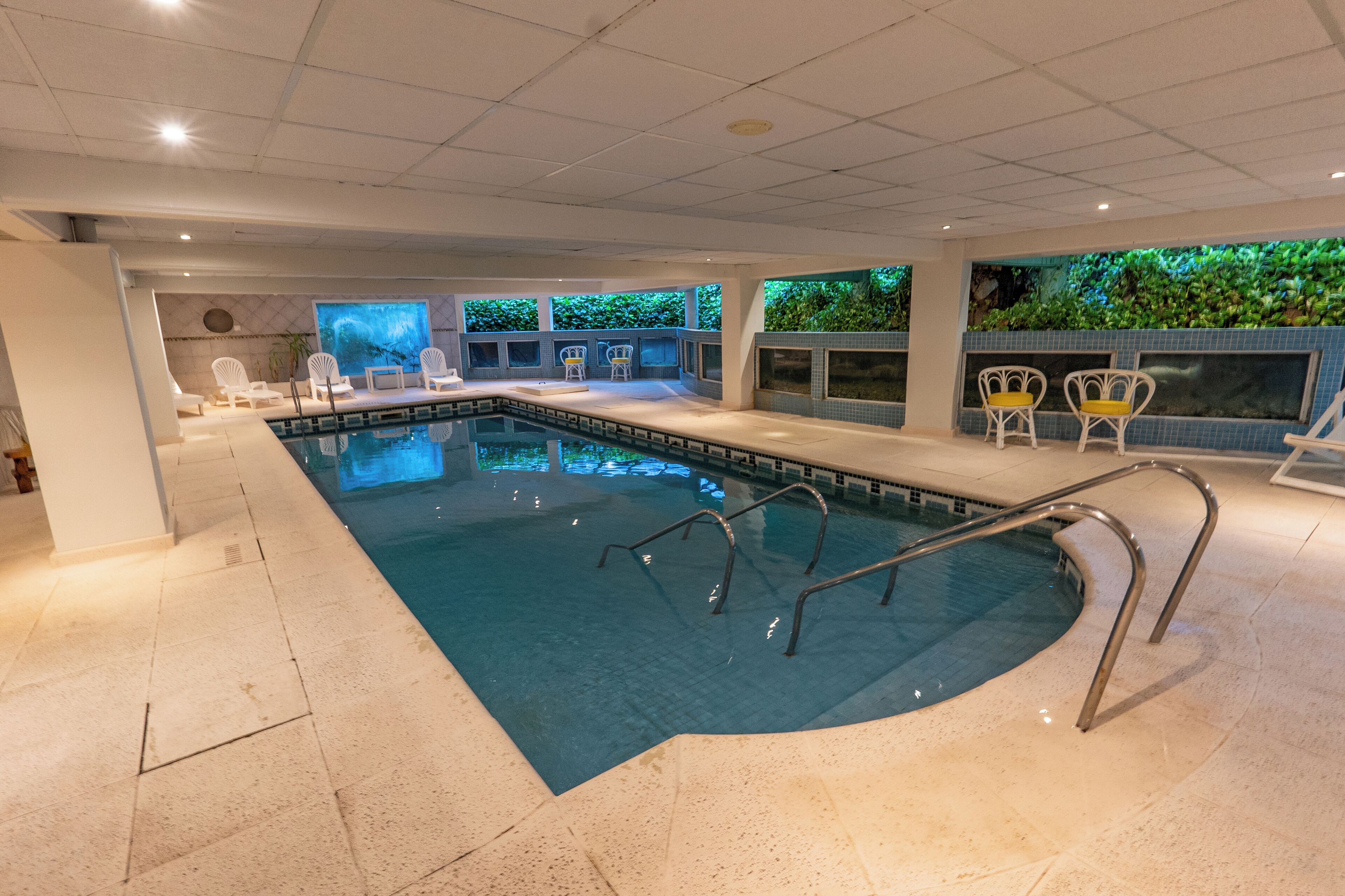 An indoor swimming pool with clear water, surrounded by white lounge chairs and a tiled floor.
