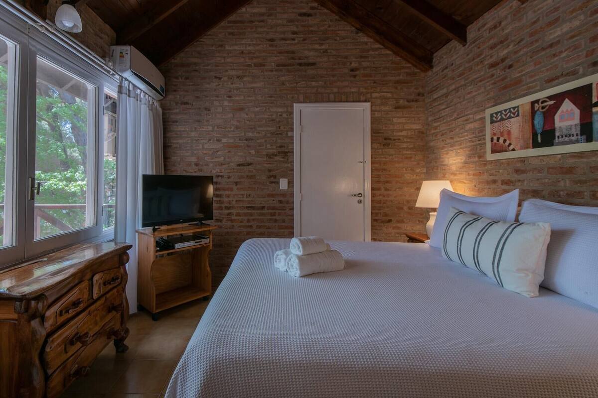 Quarto com parede de tijolos, cómoda de madeira, televisão sobre suporte, cama com almofadas e uma porta.
