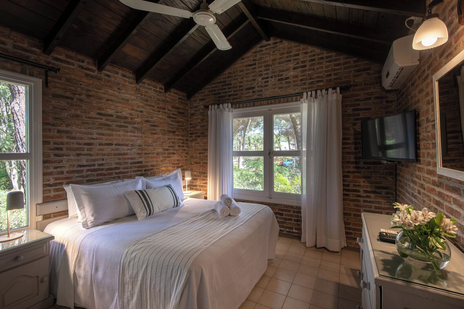 Un dormitorio con pared de ladrillo, cama con ropa blanca, ventilador de techo, ventana con cortinas, televisor montado en la pared y un florero con flores sobre una mesa.
