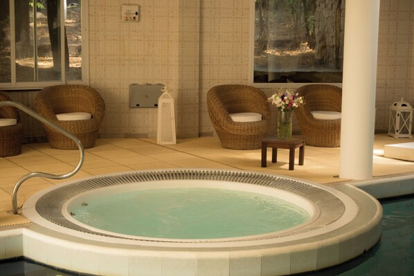 A spa area with a hot tub, wicker chairs, a small table with a vase of flowers, and a view of a natural landscape through a window.