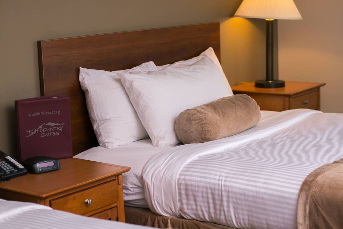 A hotel room with two beds, a nightstand with a phone and a guest directory, and a bedside lamp.