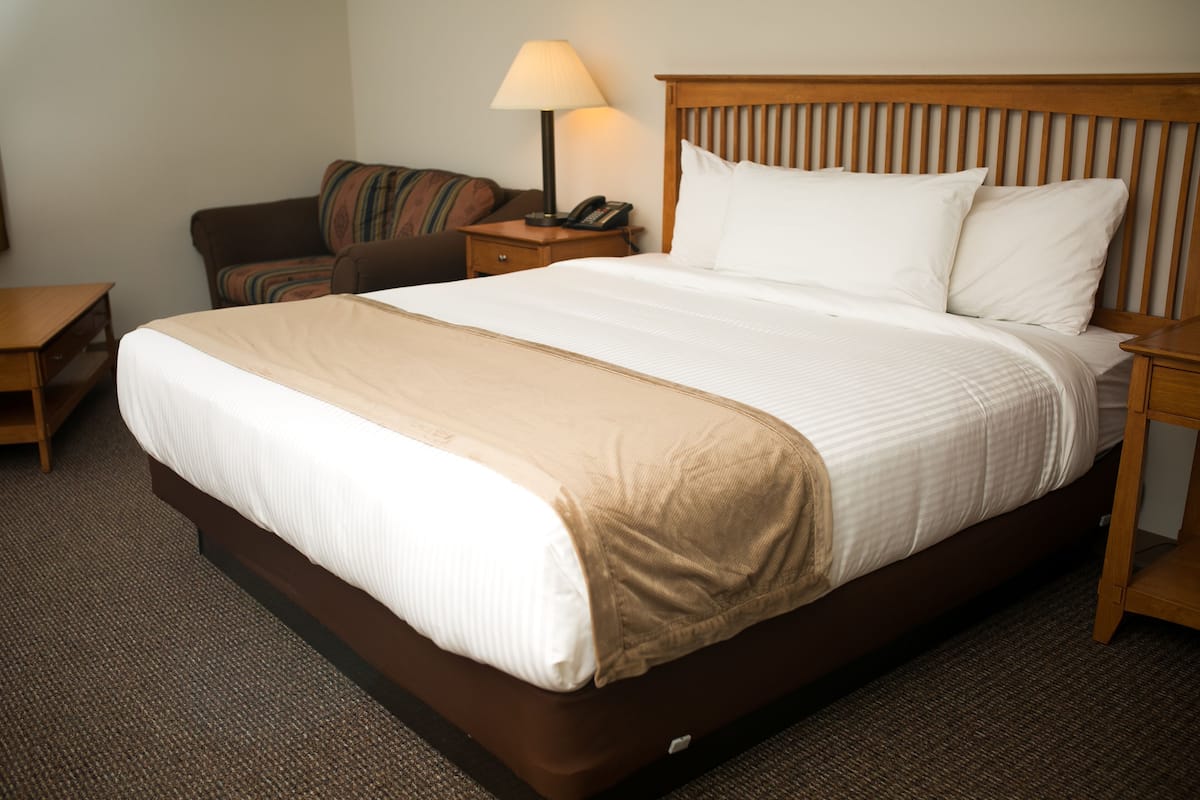 A hotel room with a large bed, a wooden headboard, a nightstand with a lamp, a telephone, and a brown armchair.