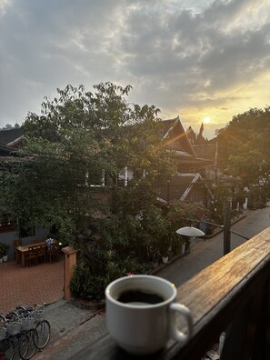 Een witte koffiekop op een houten plank met uitzicht op traditionele gebouwen en bomen onder een bewolkte lucht.
