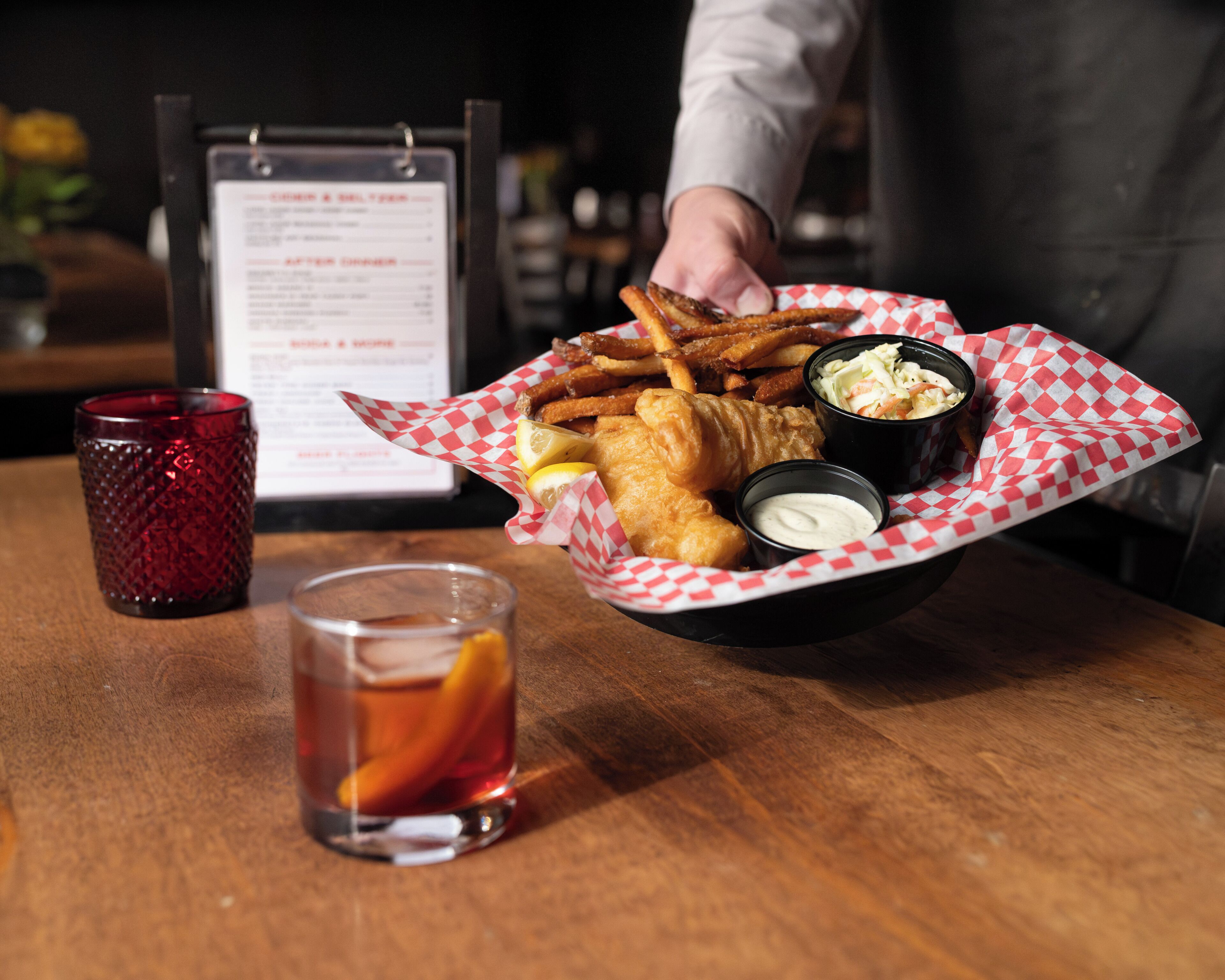 A basket of fish and chips, coleslaw, and dipping sauces on a wooden table.