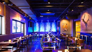 Ein Restaurant mit mehreren Tischen und Sitzecken, einer Ziegelwand mit aufgehängtem Tierkopf und blau beleuchtetem Hintergrund.