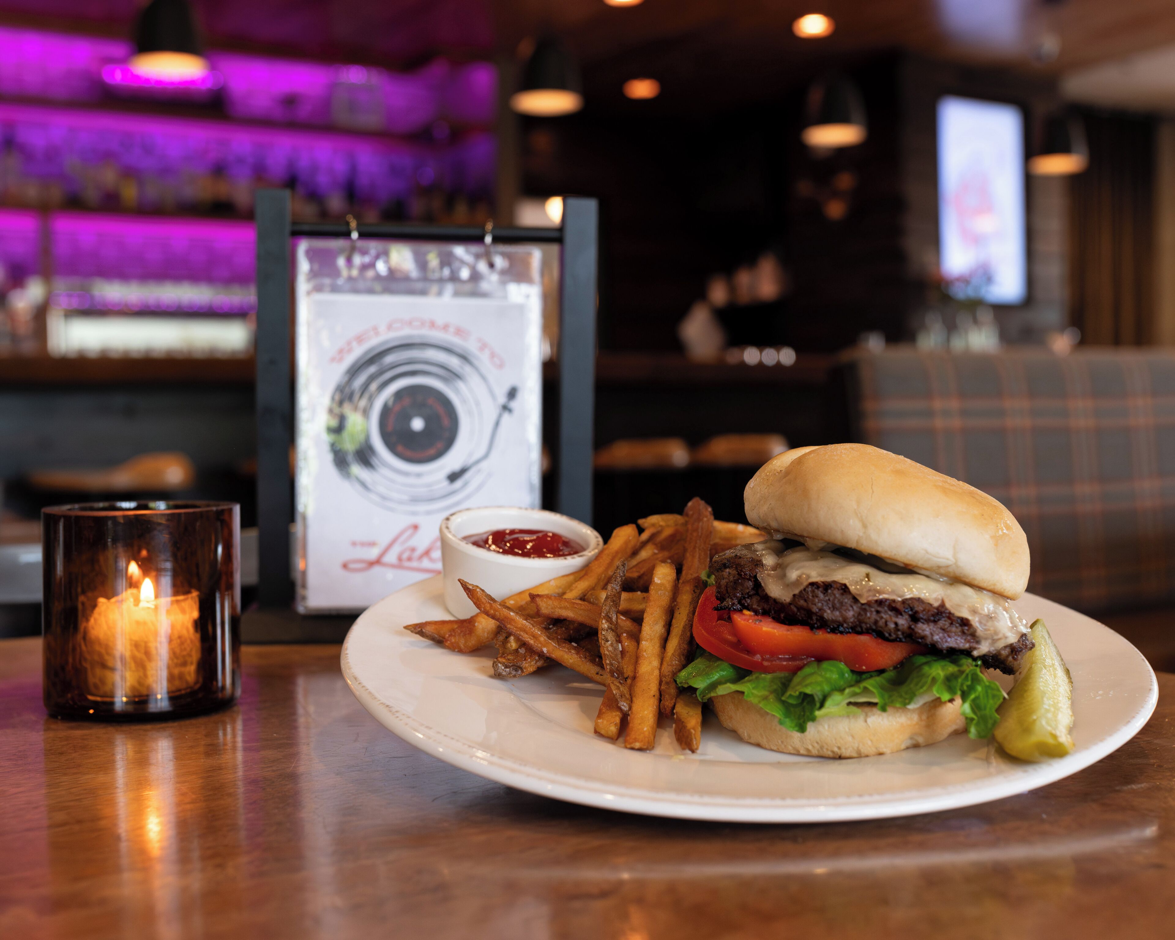 A burger with fries and ketchup on a plate, a candle, and a menu in the background.