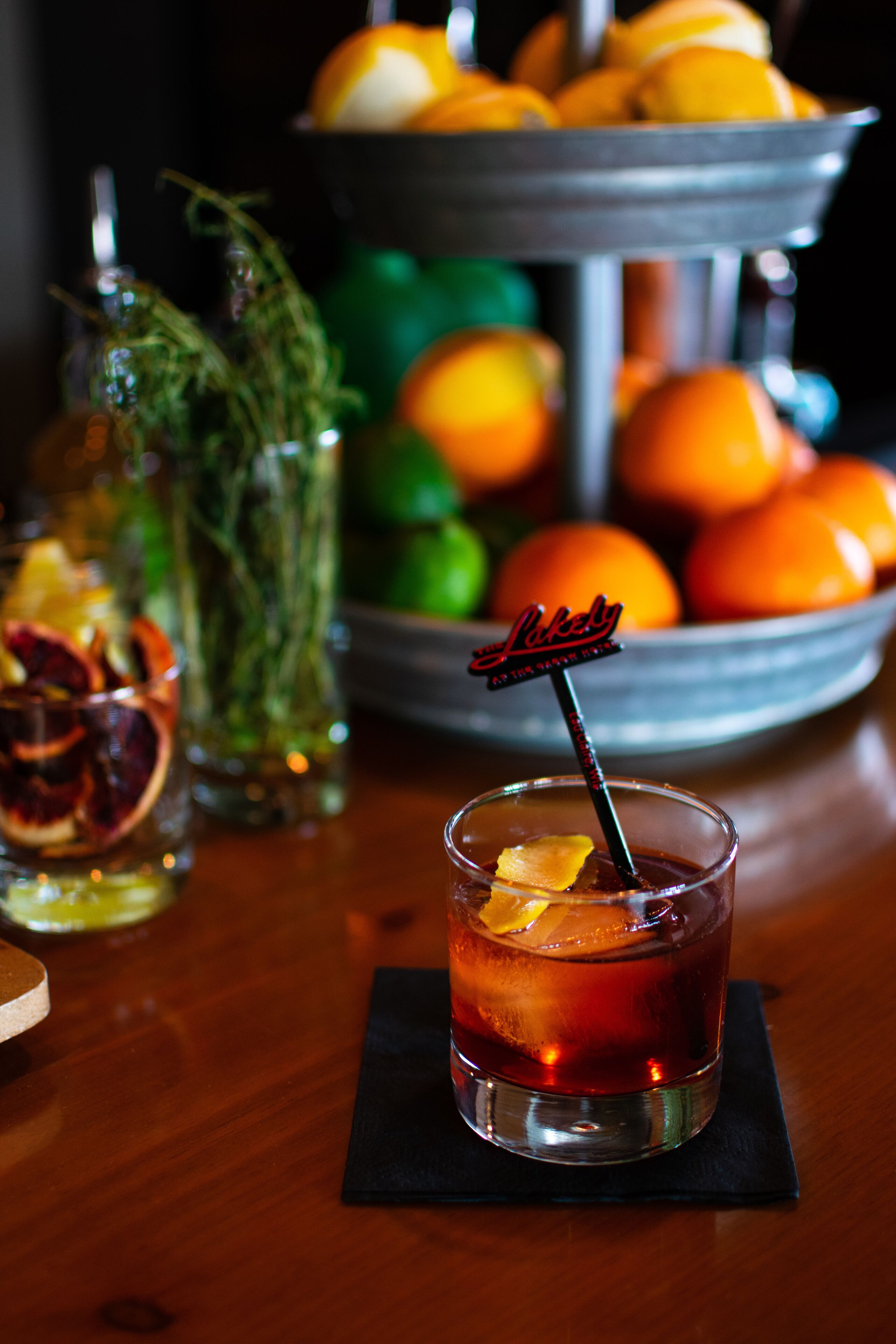A glass of red beverage with a lemon slice and a 'Likely' branded straw, placed on a black coaster. In the background, there is a metal tray with oranges and lemons, and a bottle with a green plant.
