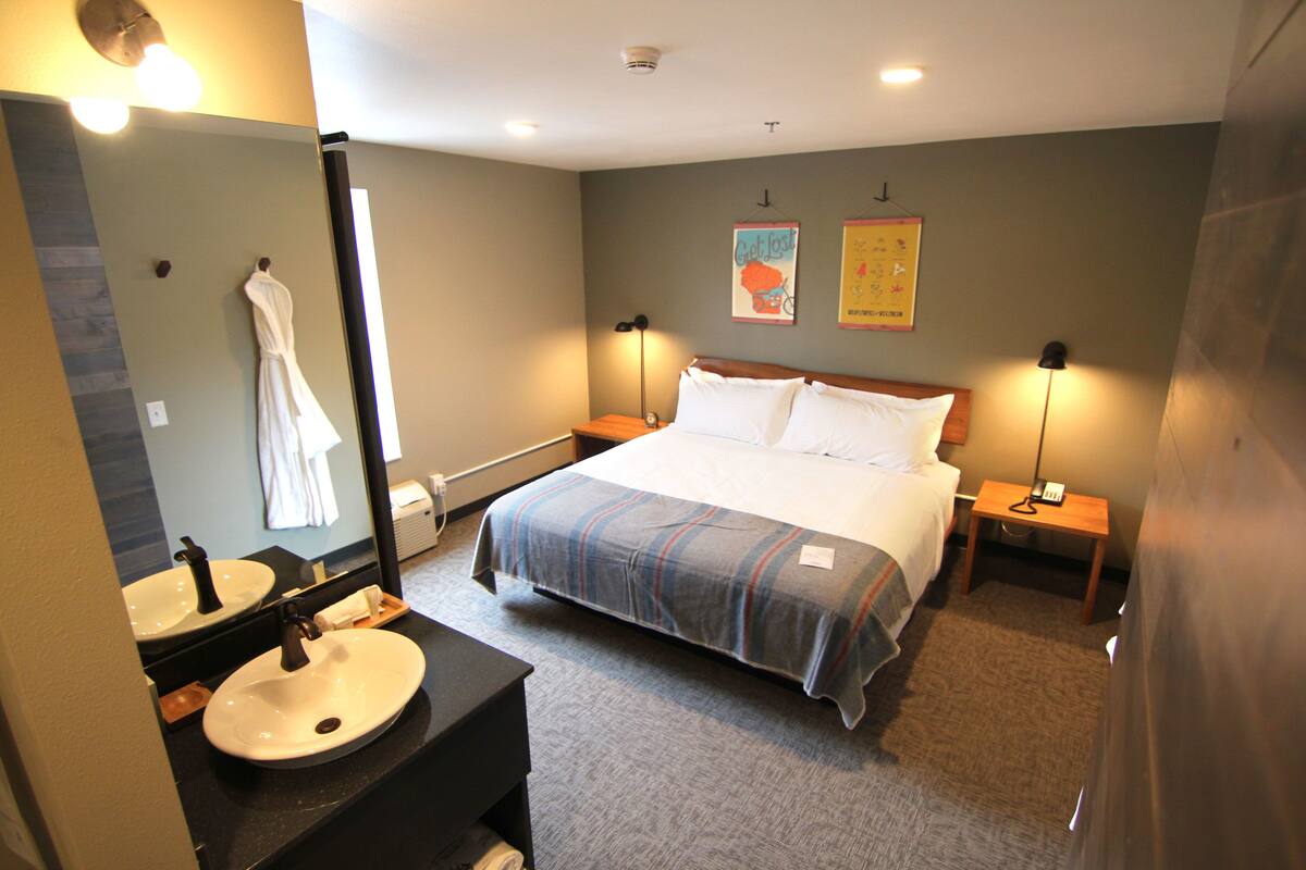 Una habitación de hotel con cama, mesitas de noche, espejo, lavamanos y toallero.