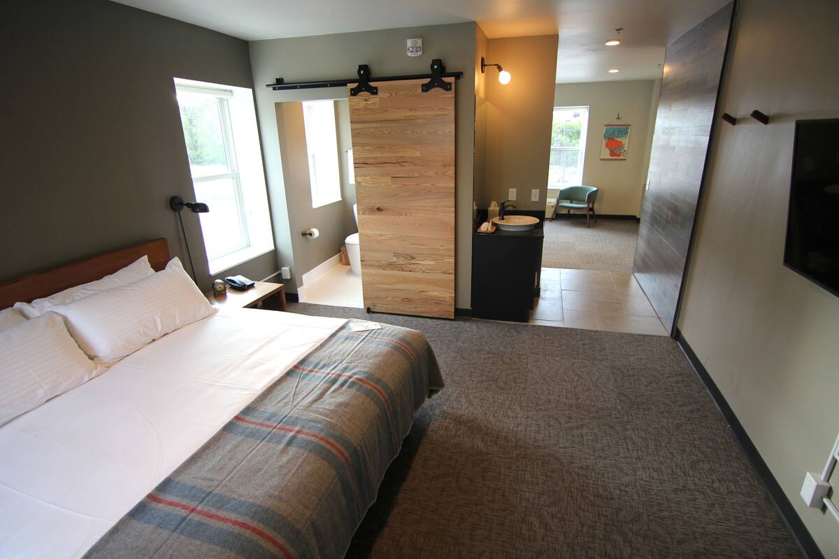 Una habitación de hotel con una cama, una puerta corredera de madera, un baño con lavamanos y retrete, y una silla.