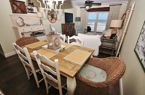 Un coin repas avec une table en bois et des chaises blanches, un panier en osier et une vue sur l’océan depuis un balcon.