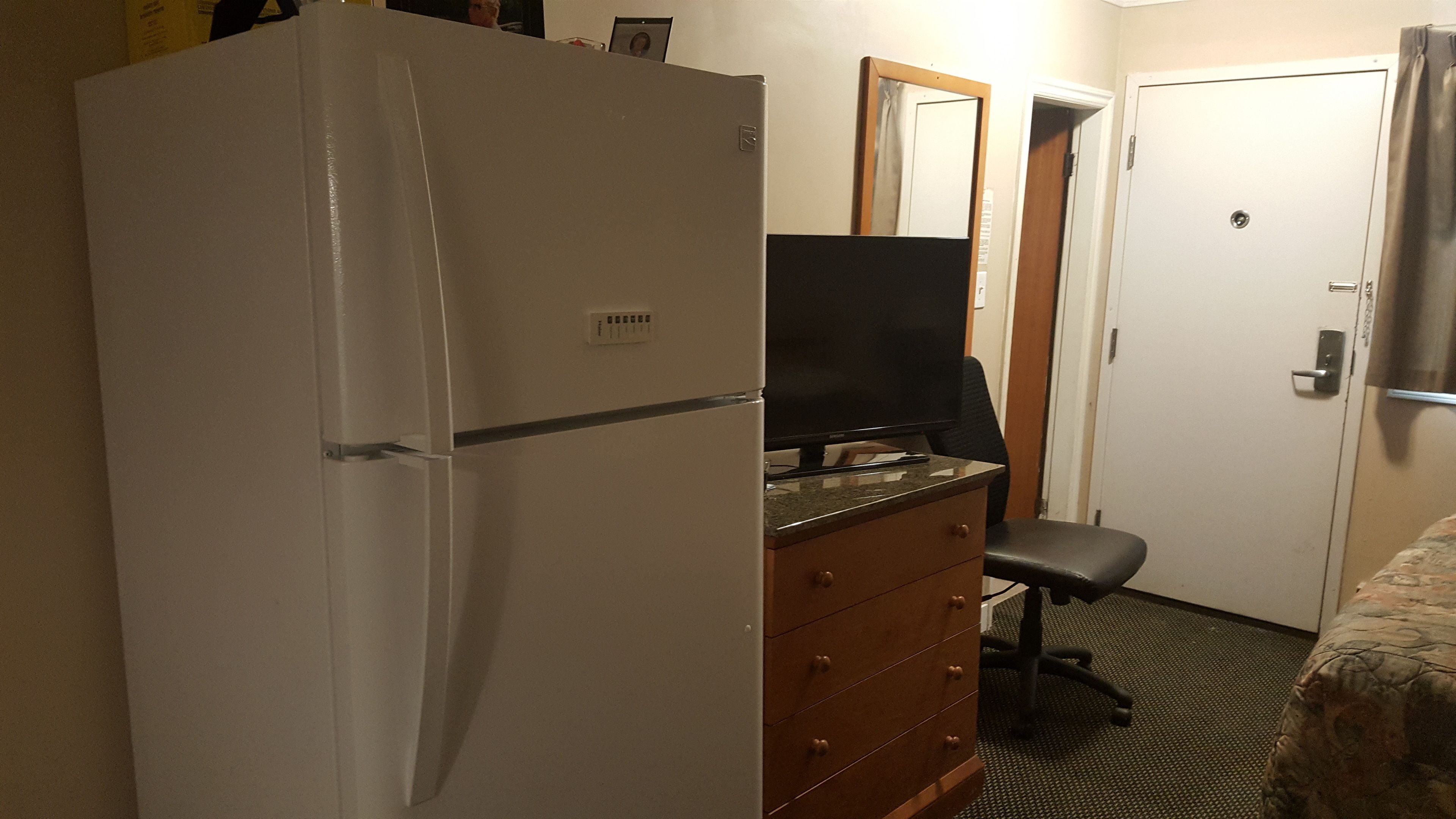 A compact hotel room with a white refrigerator, a wooden dresser, a black office chair, and a television on a small table.