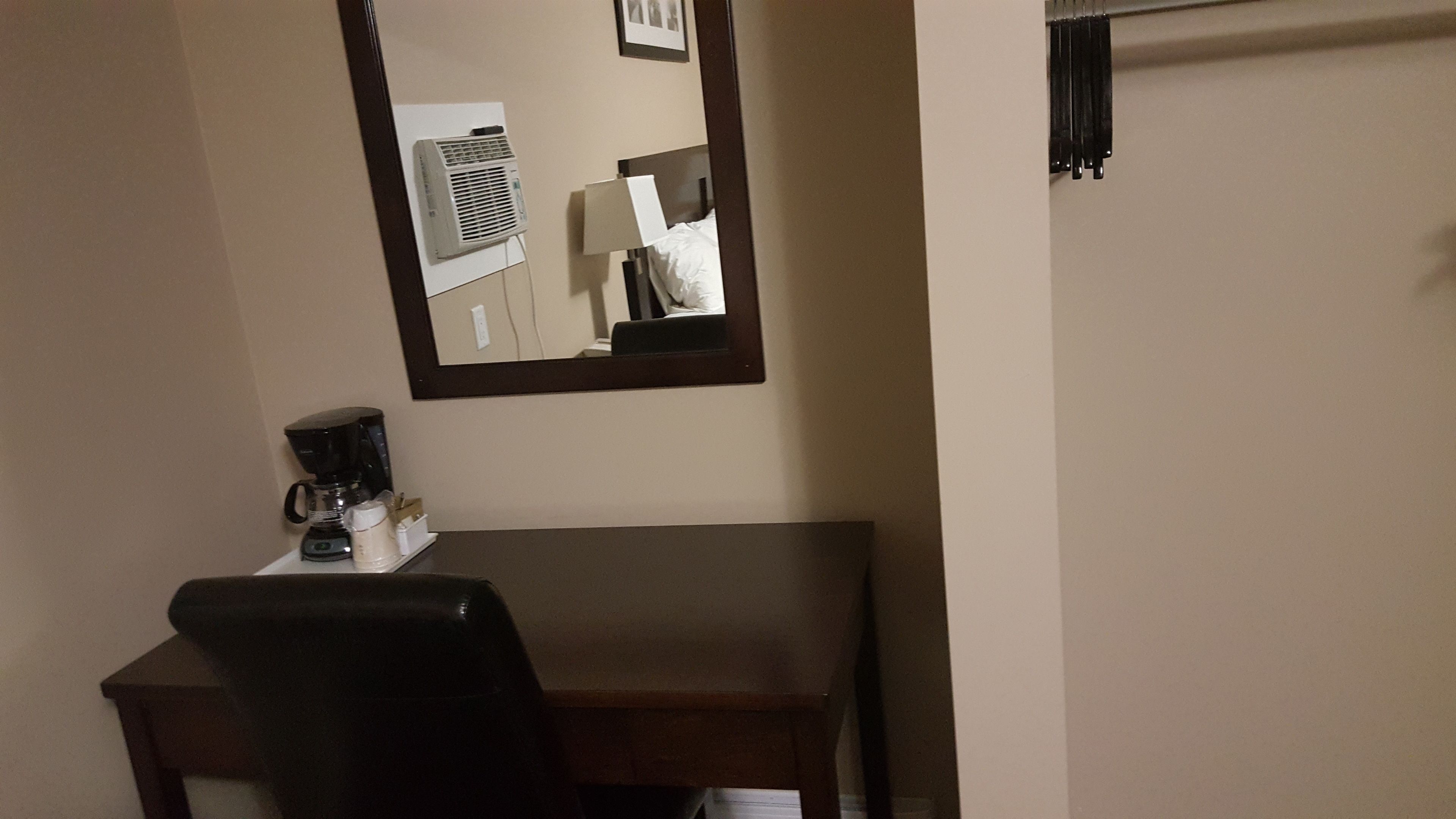 A hotel room with a desk, a chair, a coffee maker, a mirror, and a wall-mounted air conditioner.