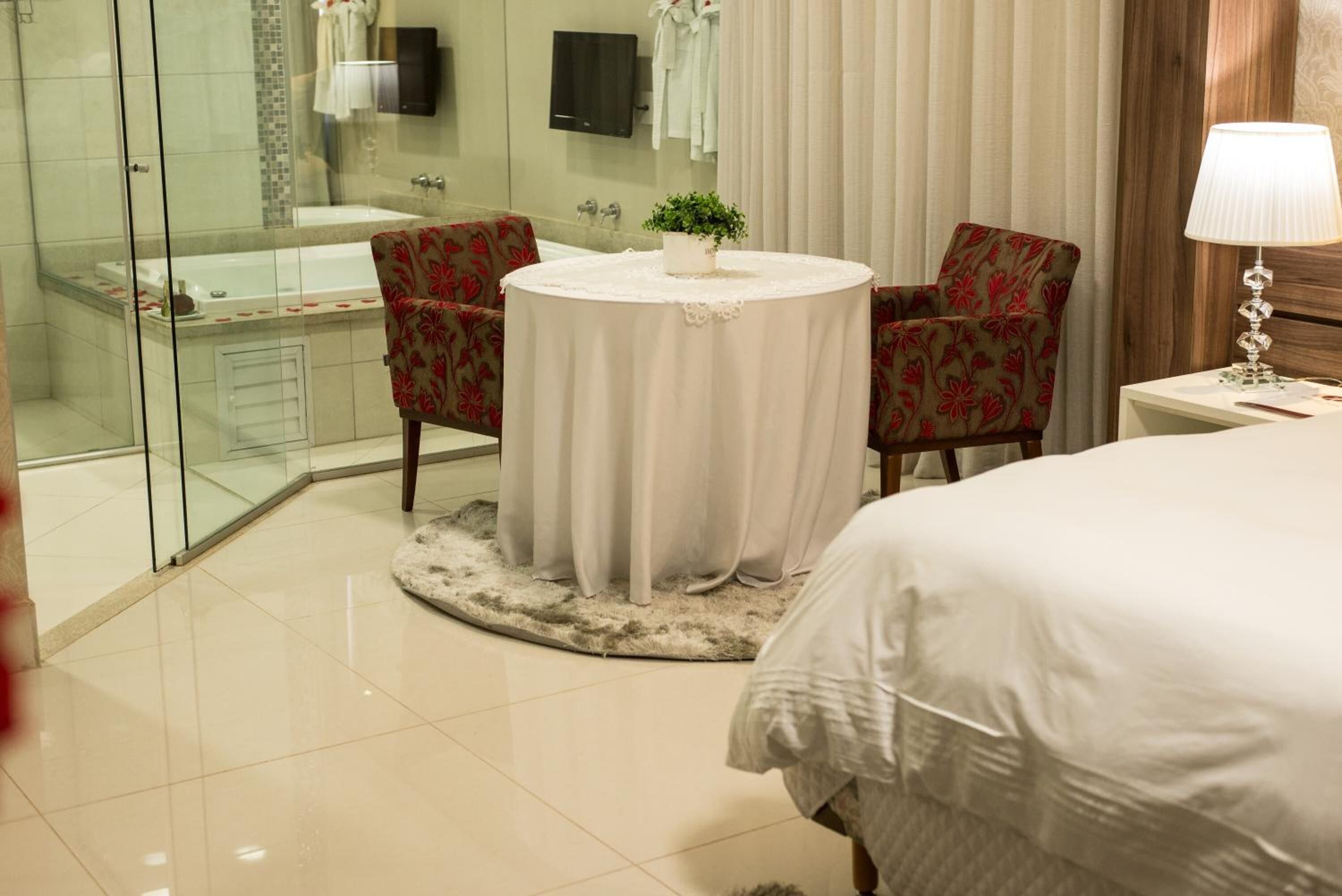 A hotel room with a glass-enclosed shower, a round table with a potted plant, two floral armchairs, a bed with white bedding, and a bedside lamp.
