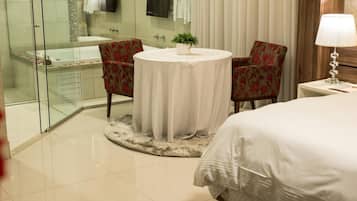 A hotel room with a glass-enclosed shower, a round table with a potted plant, two floral armchairs, a bed with white bedding, and a bedside lamp.