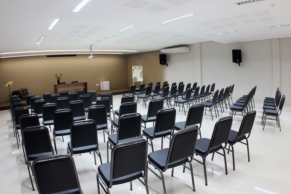 Un salón de conferencias con filas de sillas negras, un atril, una pizarra y un altavoz en la pared.