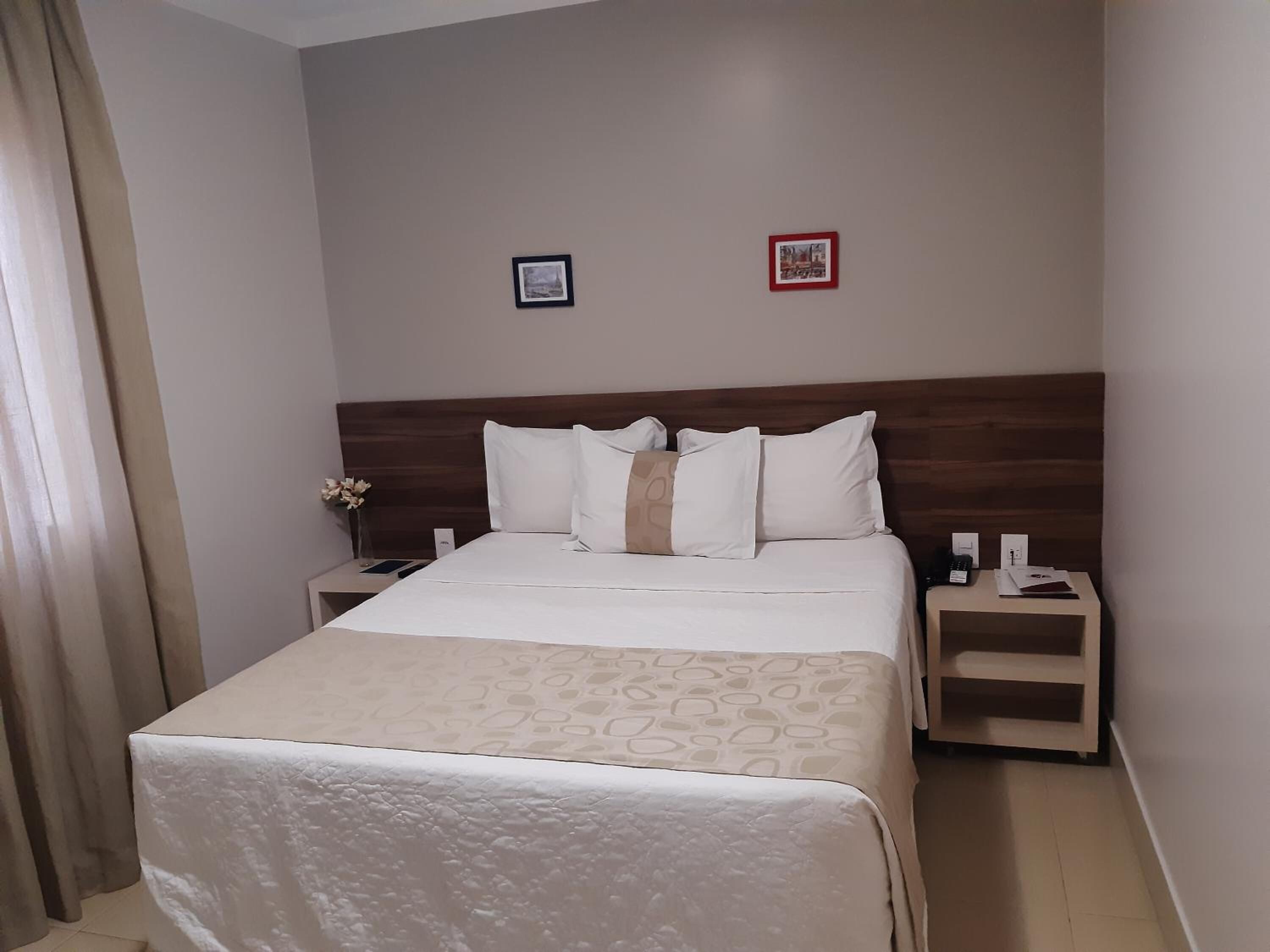 Habitación de hotel con una cama, dos mesitas de noche, una lámpara y dos cuadros en la pared.