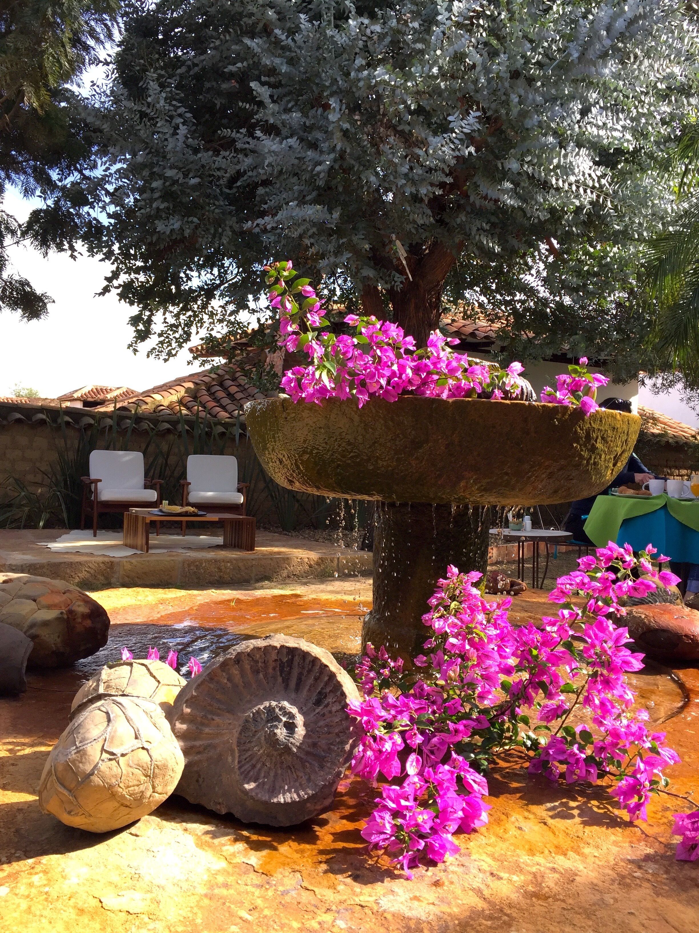 A fountain with a tree and pink flowers, surrounded by stone sculptures and a seating area with chairs and a table.