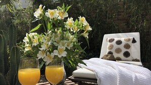 Una mesa de madera con dos vasos de jugo de naranja, un florero con flores blancas y una cama blanca con un cojín estampado.