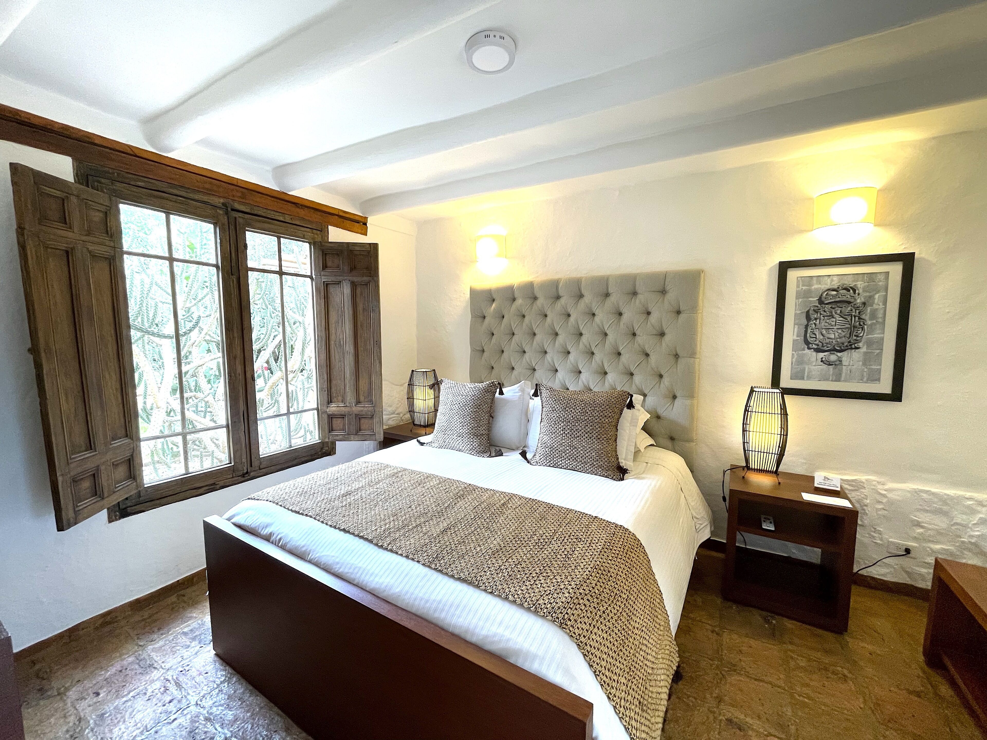 Ein Schlafzimmer mit einem großen Bett, Holzfensterrahmen, einem gebuschten Kopfteil und einem gerahmten Bild an der Wand.