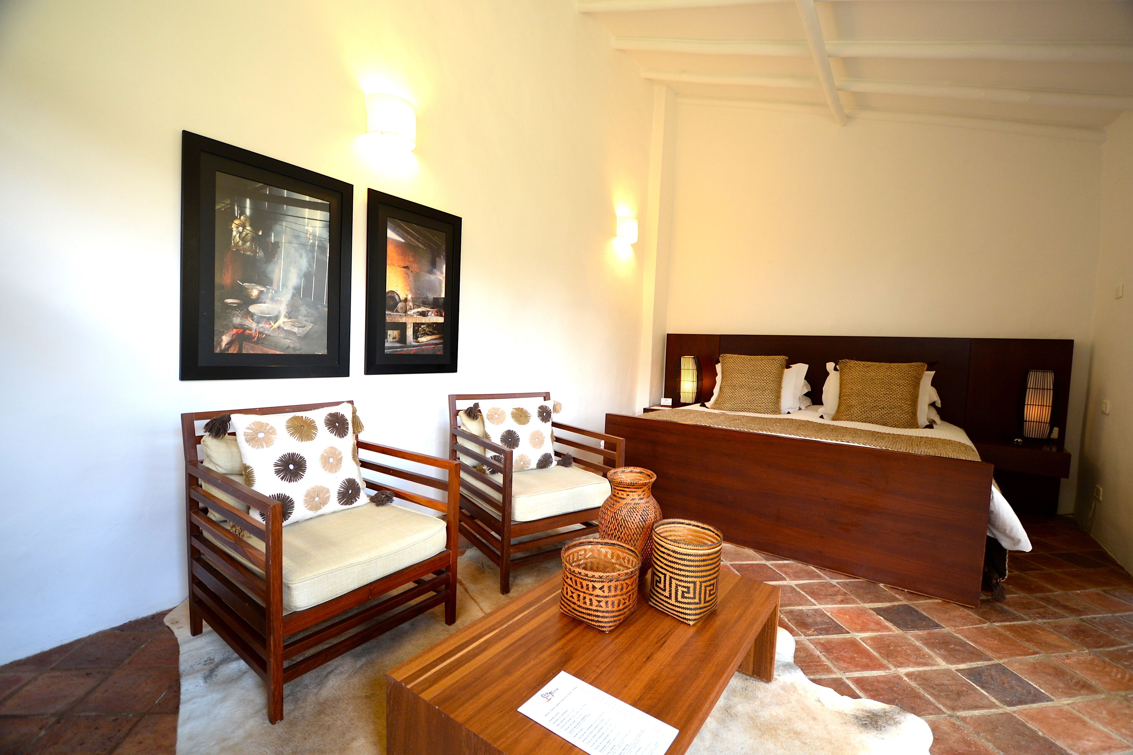A bedroom with a wooden bed, two wooden chairs with patterned cushions, a small wooden table with woven baskets, and two framed pictures on the wall.