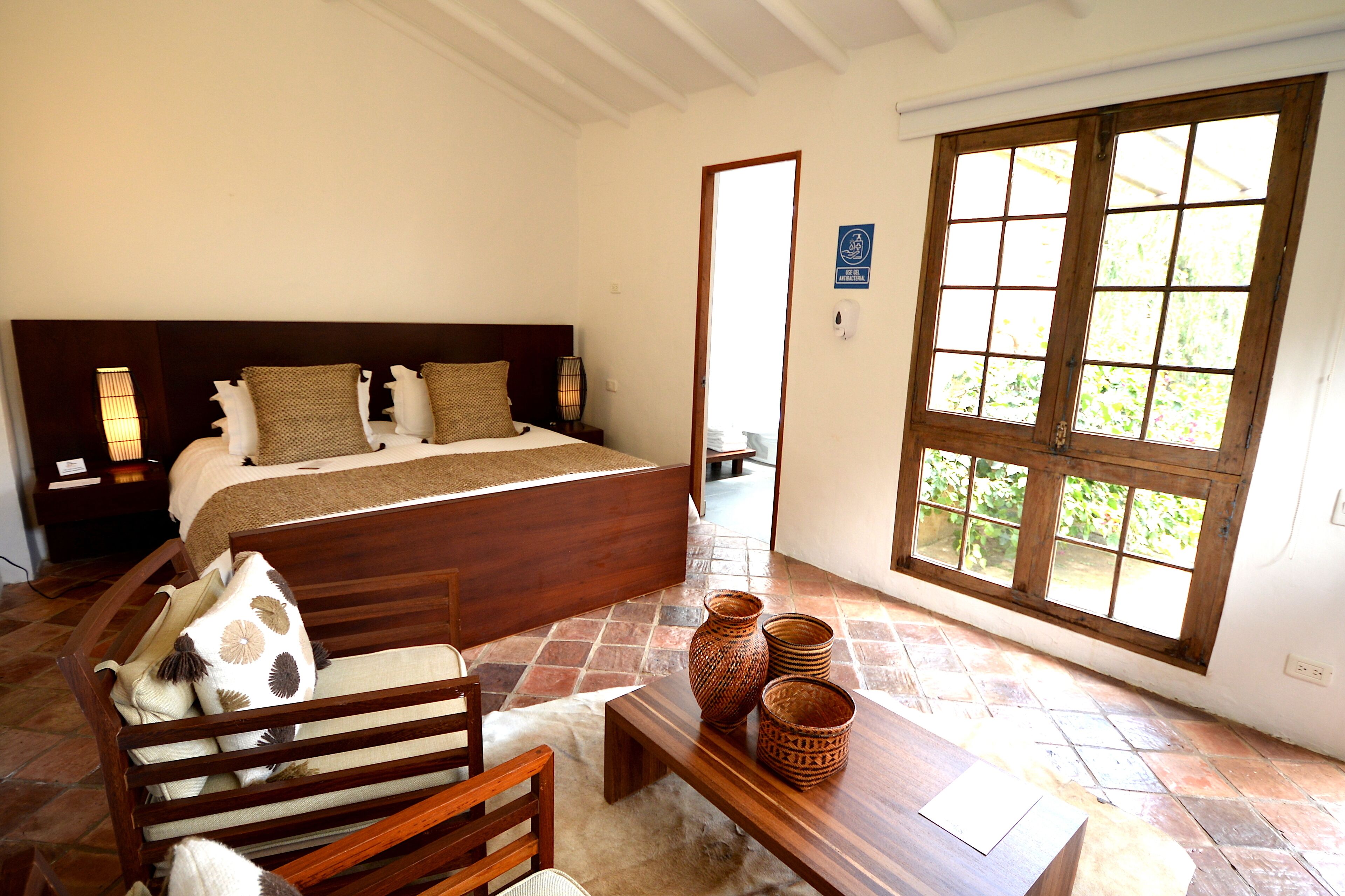 Un dormitorio con una cama grande, cabecera de madera y ventana con vistas a zonas verdes. Hay un banco de madera y una mesa con cestas decorativas.