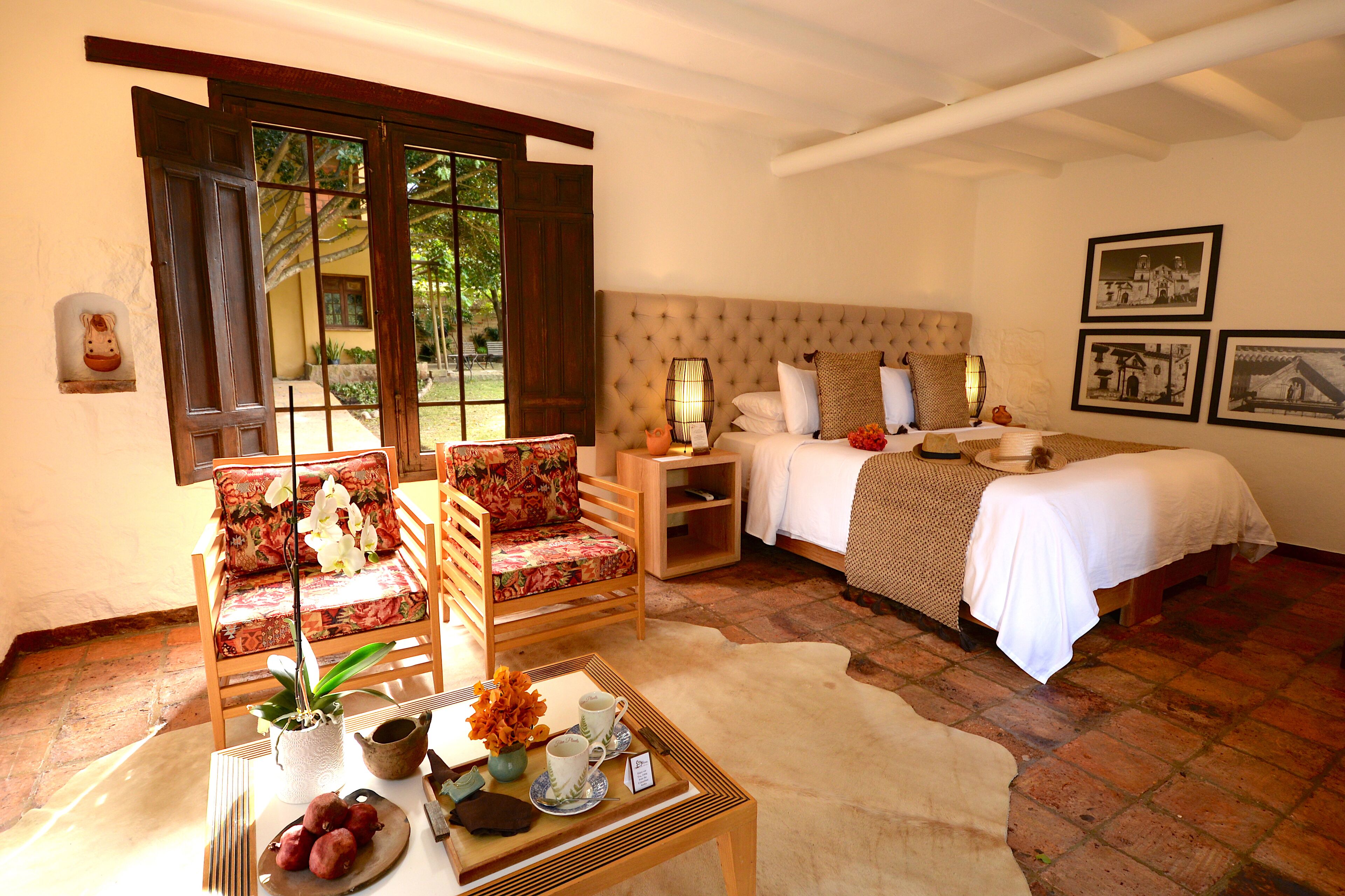 A bedroom with a bed, two chairs, a nightstand, a table with a vase and fruit, and a view of the outdoors through large windows.