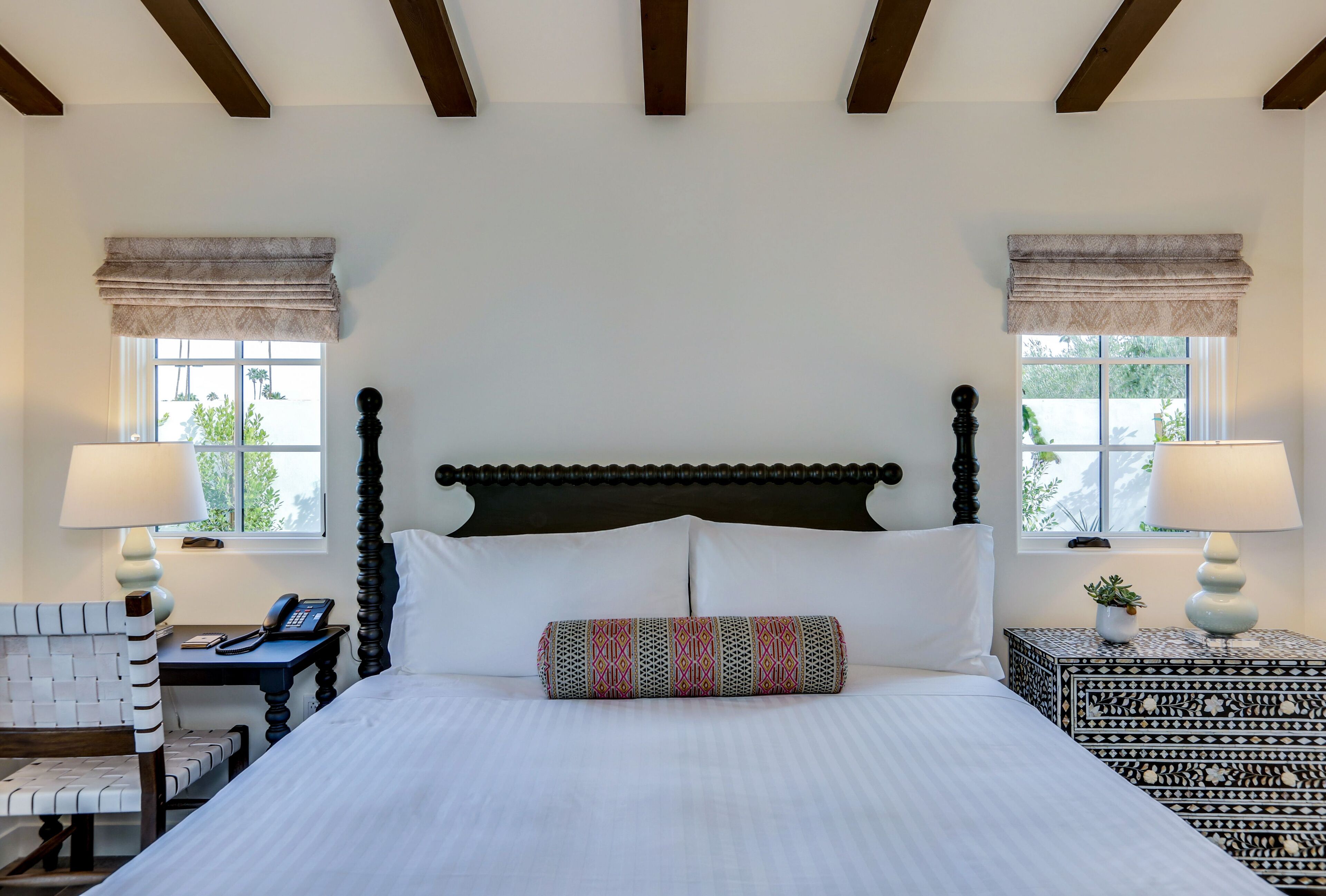 A neatly made bed with a patterned pillow, a bedside table with a lamp, and a chair with a telephone.