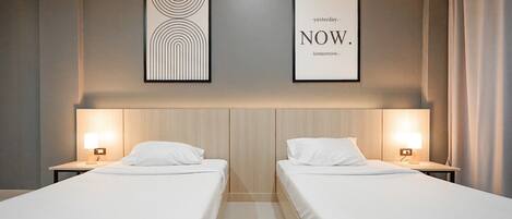 A hotel room with two beds, a headboard with a light wood finish, and two bedside tables with lamps. There are two framed artworks on the wall.