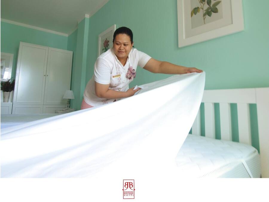 A person adjusting a bedspread in a room with a white wardrobe, a framed picture of a flower on the wall, and a logo for Boonsiri Boutique Hotel.