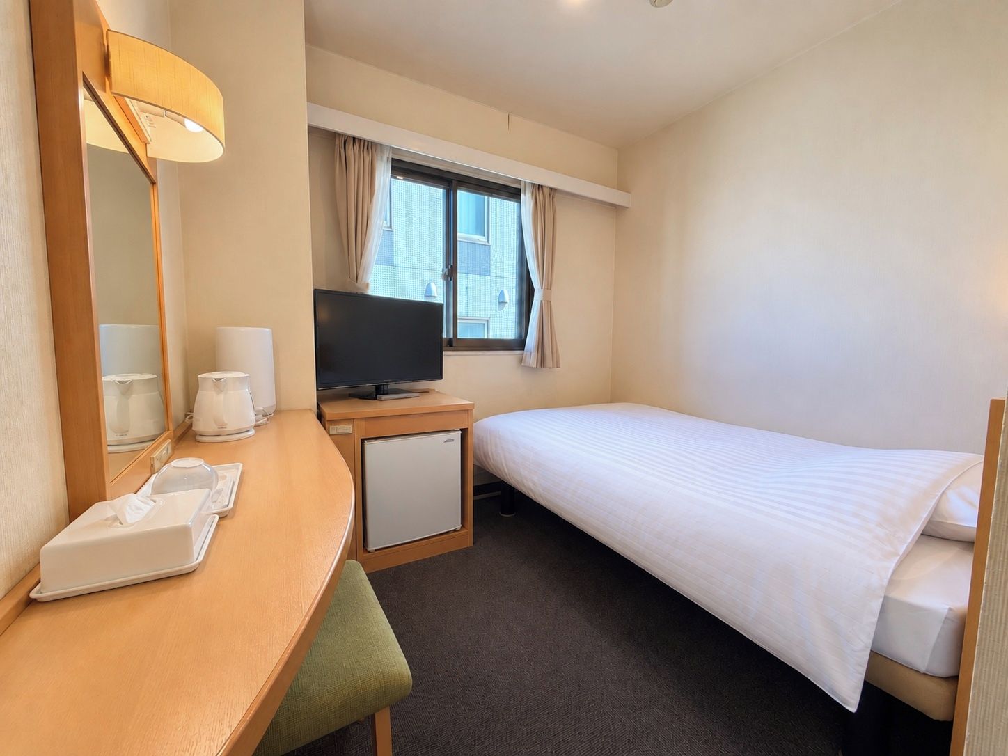Quarto de hotel com uma cama de solteiro, uma escrivaninha com televisão, um frigorífico pequeno, um espelho e uma janela com cortinas.