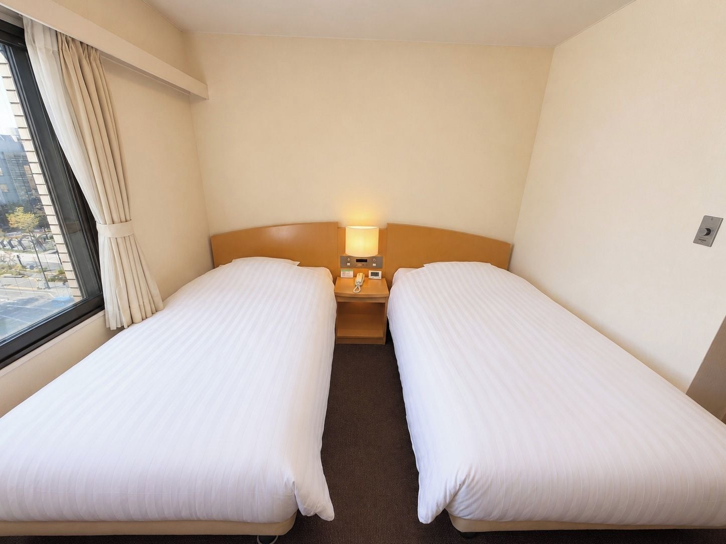 A hotel room with two single beds, a bedside table with a lamp, and a window with curtains.