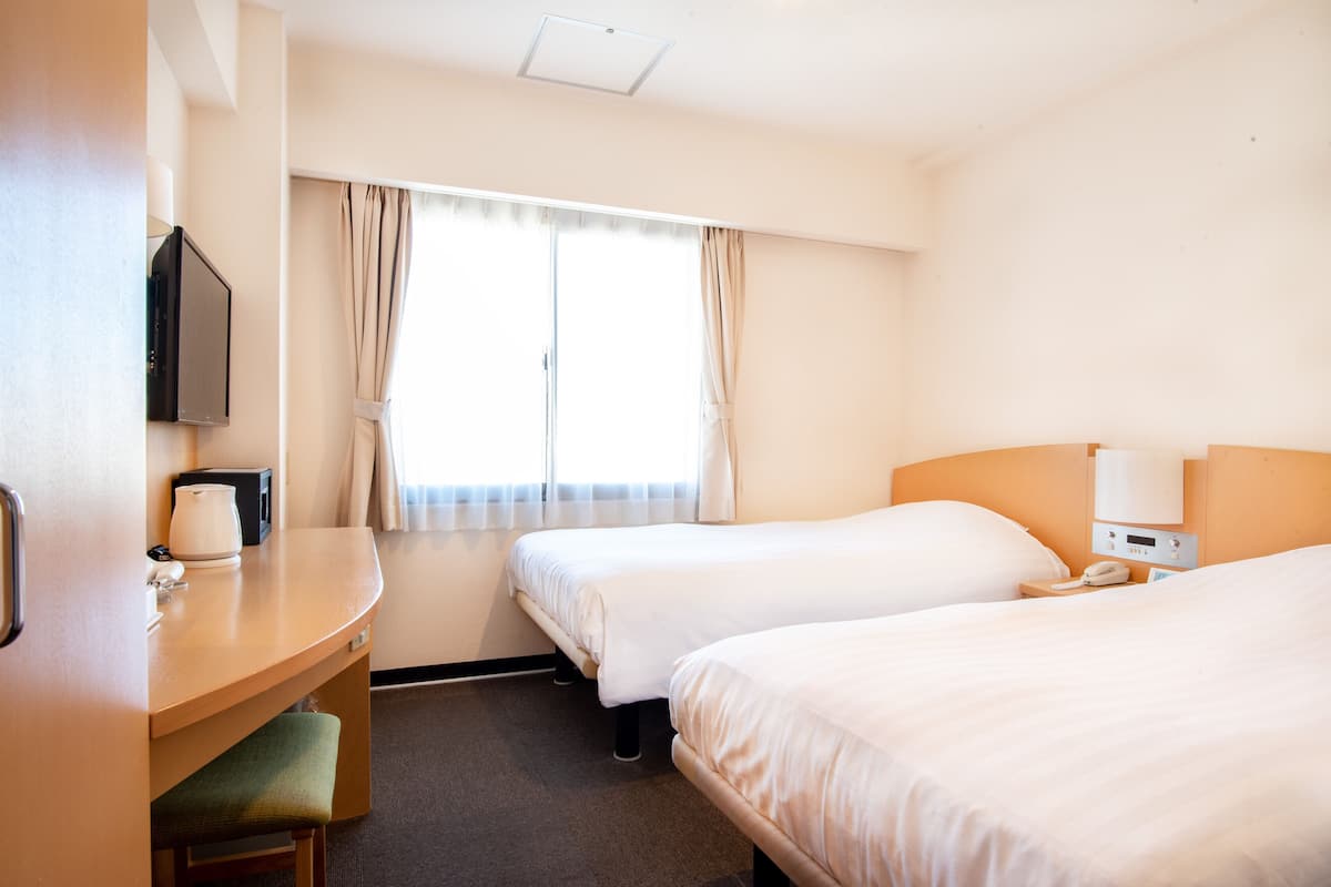 A hotel room with two beds, a desk with a chair, a TV, and a window with curtains.