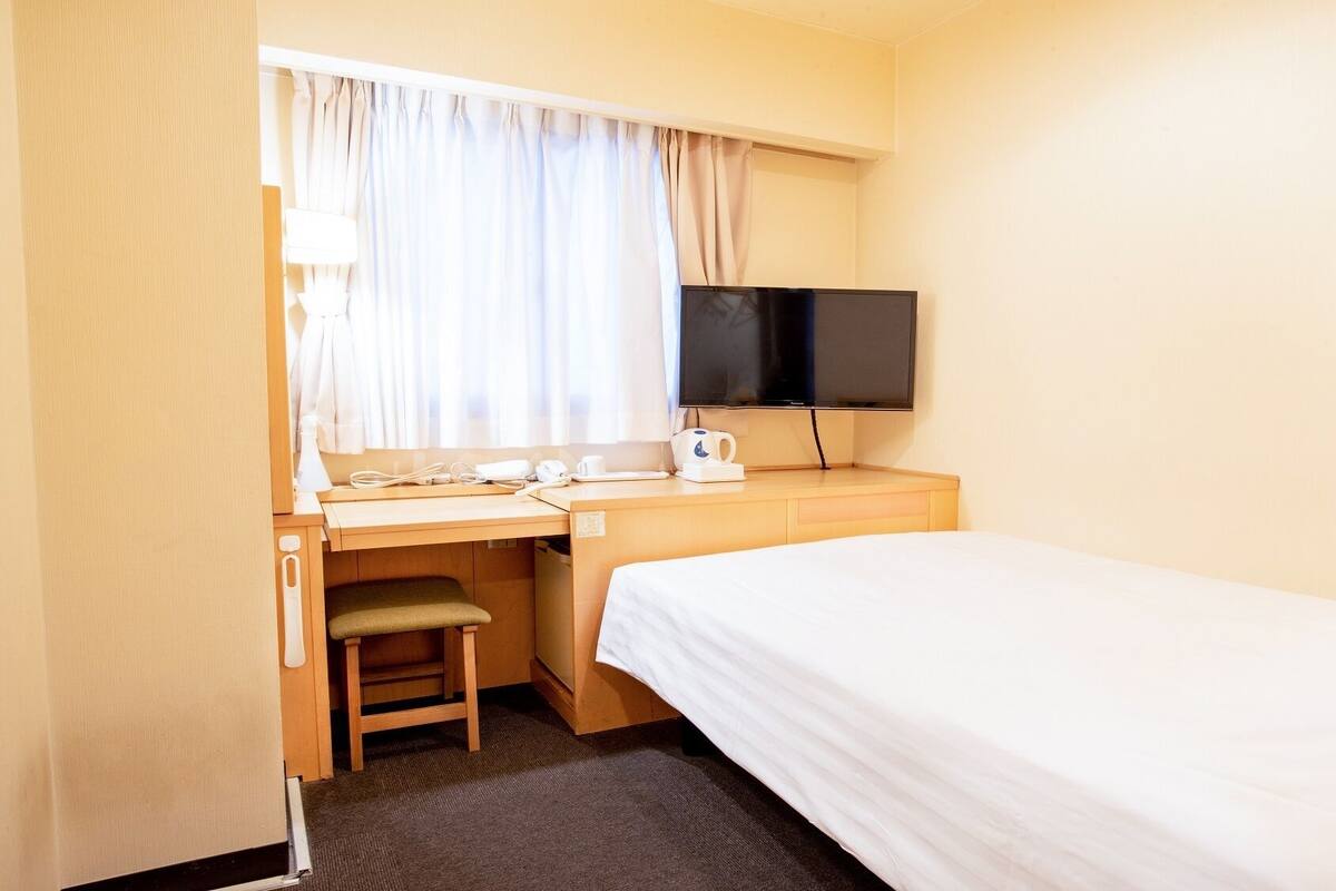A hotel room with a bed, a desk with a chair, a television, and a window with curtains.