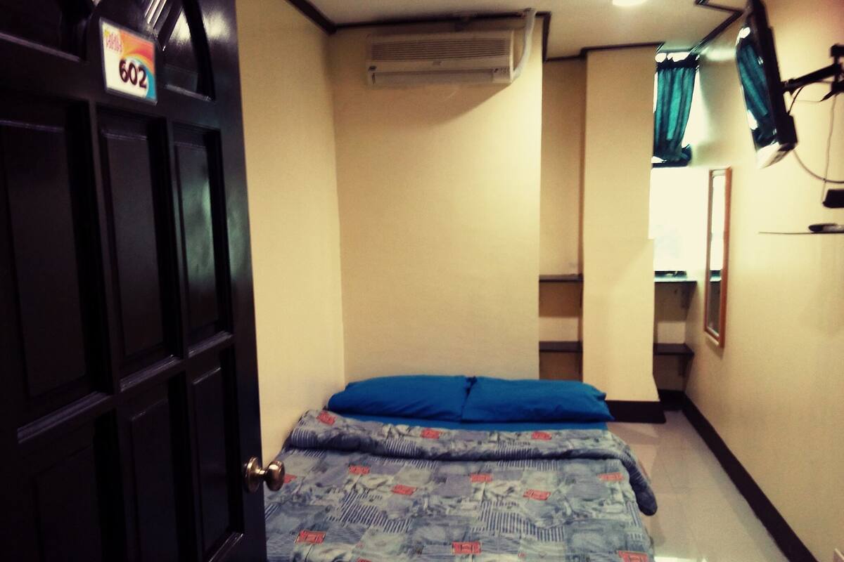 A room with a bed, a door with a number sign, an air conditioning unit, and a mirror on the wall.