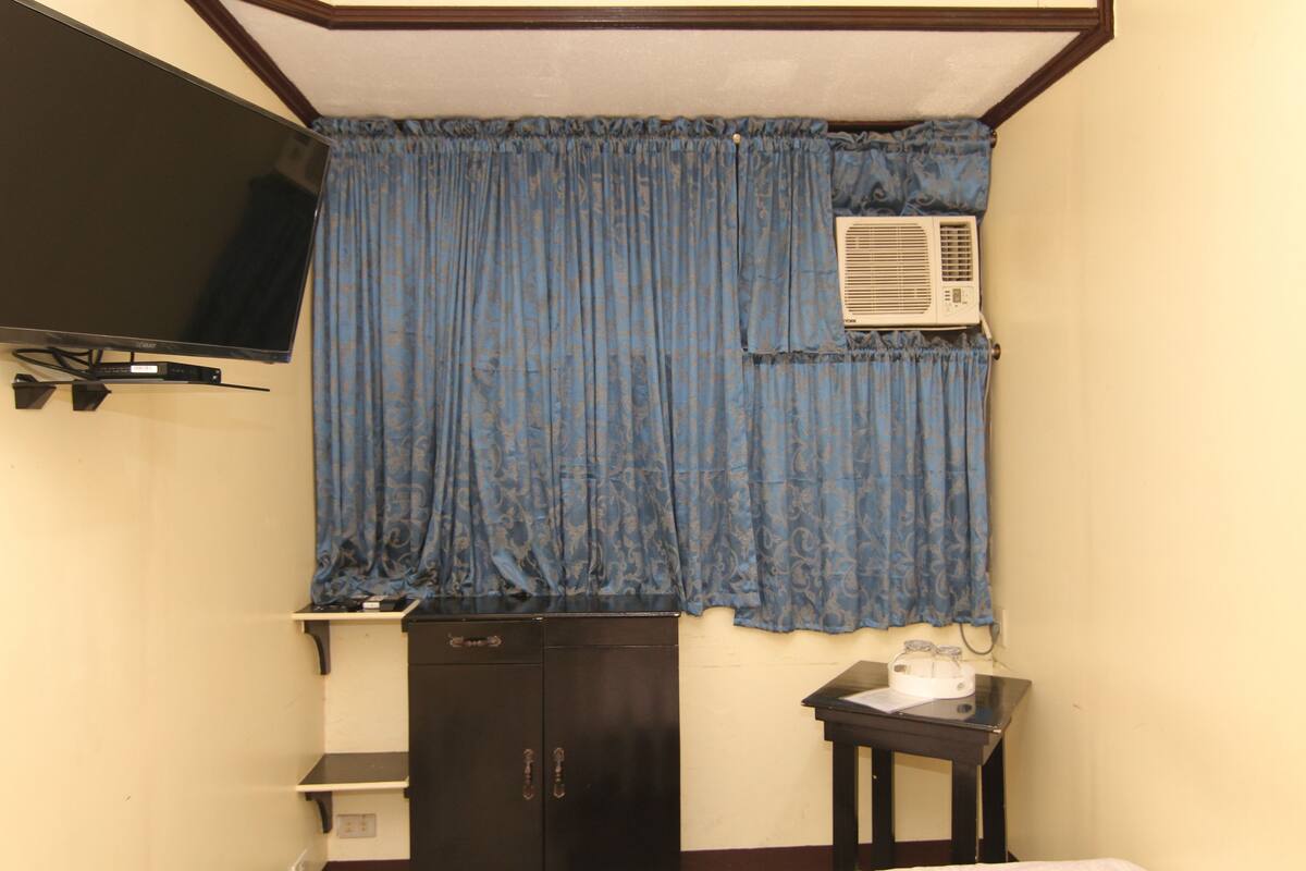 A room with a flat-screen TV mounted on the wall, a window with blue curtains, a small table with a white object, and a dark-colored wardrobe.