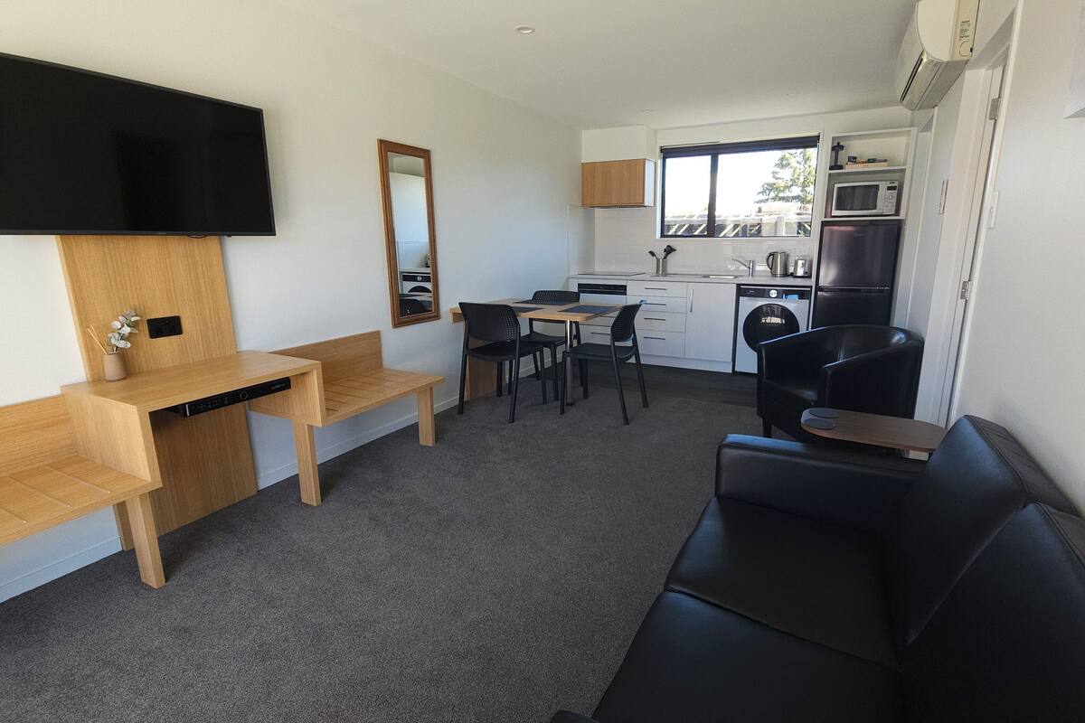 A compact living space with a flat-screen TV, a wooden desk, a black leather couch, a dining table with four chairs, a small kitchen area with a microwave and refrigerator, and a window providing natural light.
