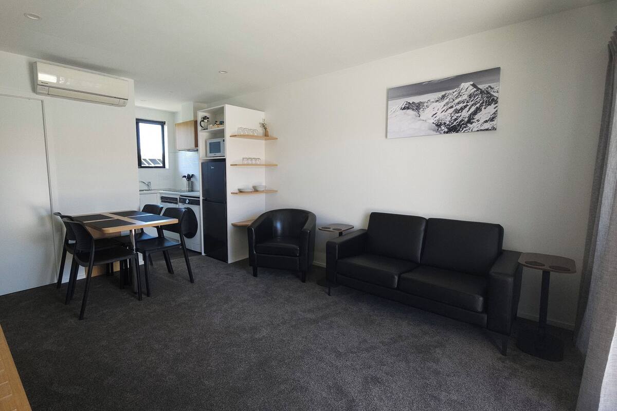 A compact living space with a kitchenette, a dining area, a black leather sofa, and a mounted artwork.