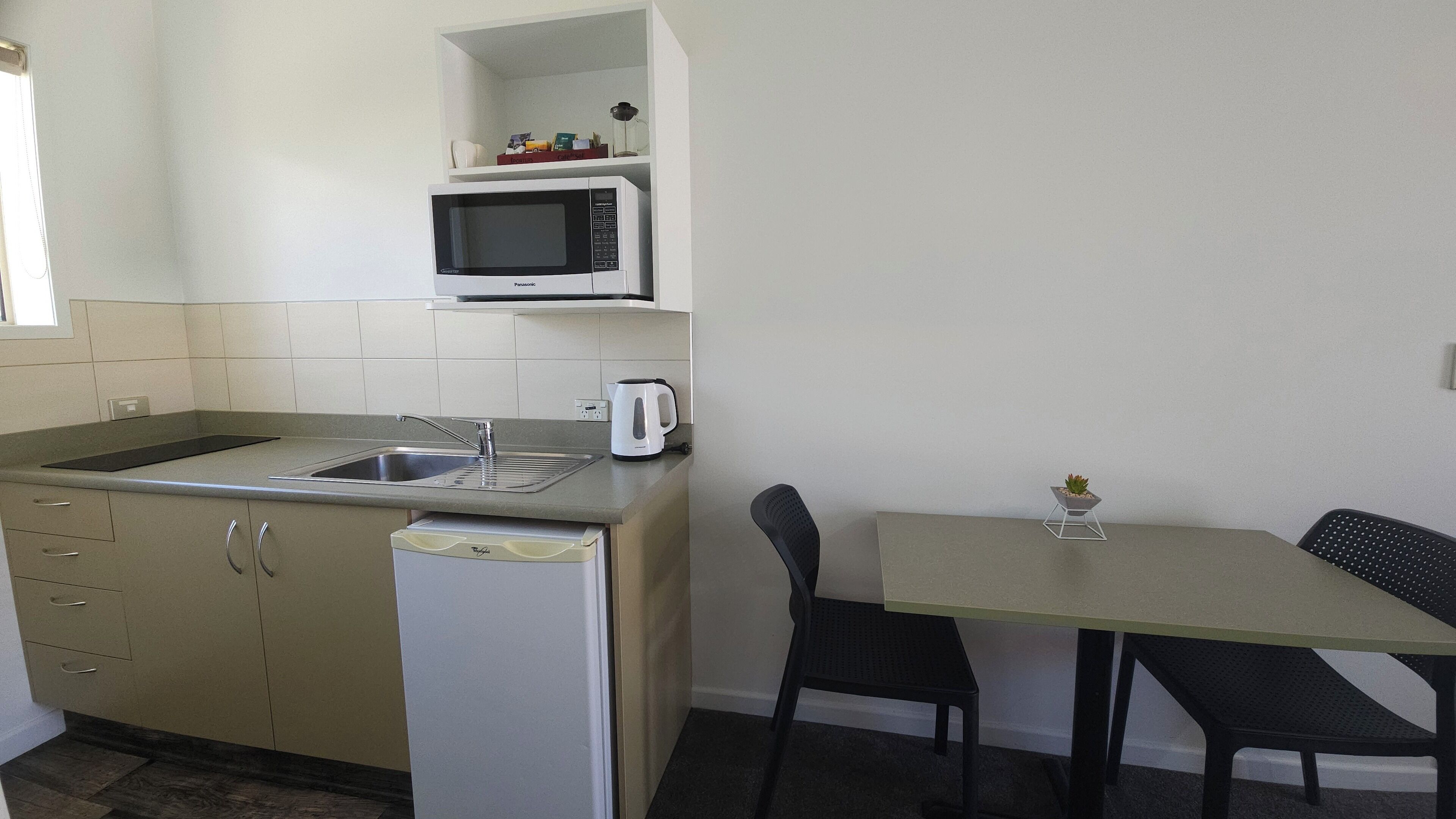A compact kitchen with a microwave, sink, and a small refrigerator. There is a dining table with two chairs and a small potted plant on the table.