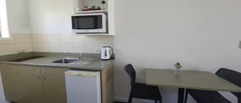 A compact kitchen with a microwave, sink, and a small refrigerator. There is a dining table with two chairs and a small potted plant on the table.