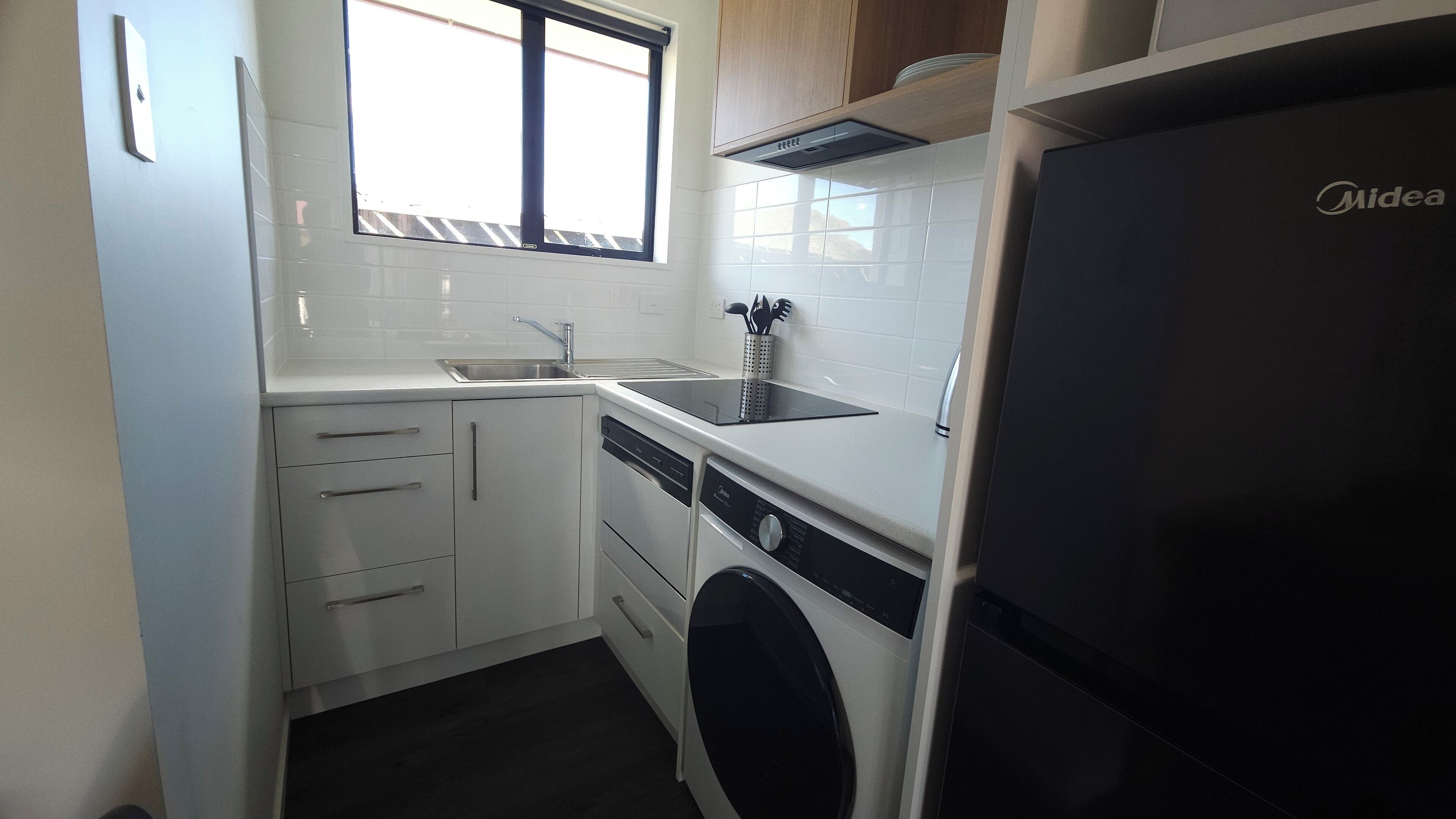 Una cocina compacta con armarios blancos, lavavajillas integrado, refrigerador negro y una ventana que proporciona luz natural.