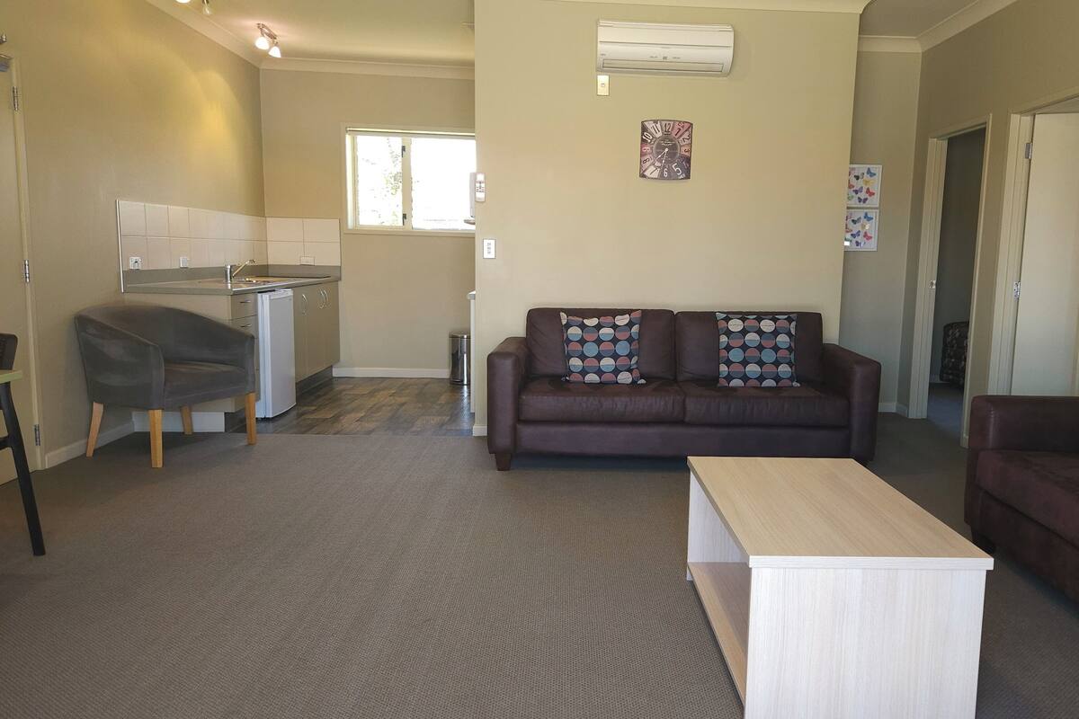A living room with a brown sofa, a wooden coffee table, and a kitchen area with a dishwasher and a window.
