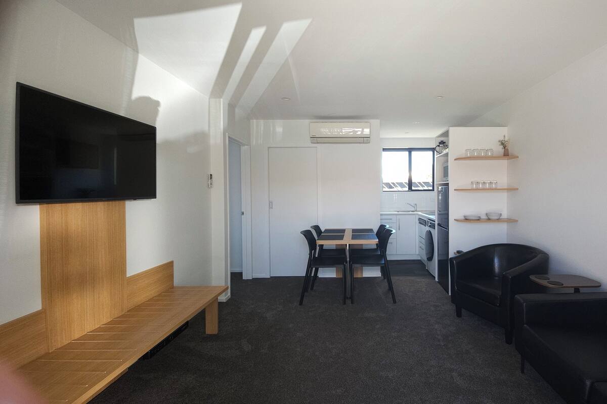 A modern living room with a flat-screen TV, a wooden bench, a dining table with chairs, a black leather sofa, and a kitchen area with shelves and a refrigerator.