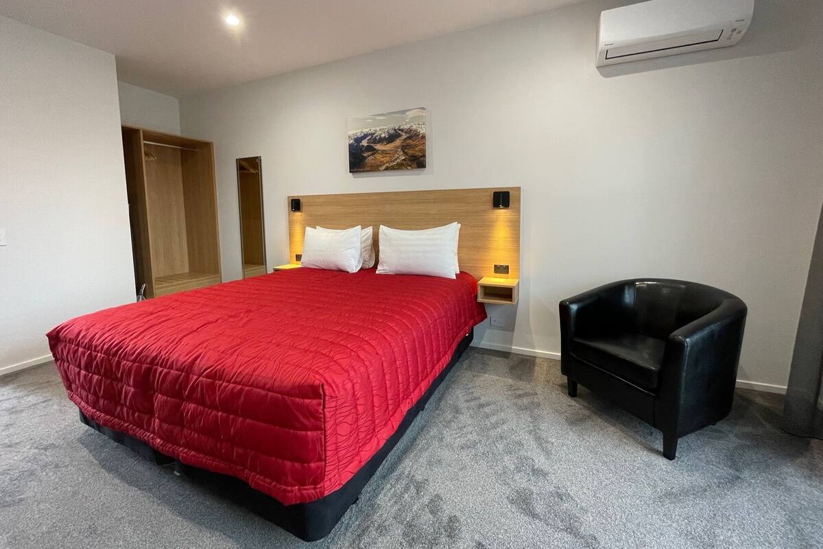 A hotel room with a large bed, a nightstand, a black leather chair, and an air conditioning unit on the wall.