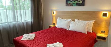 A hotel room with a large bed, red bedding, white pillows, and a painting on the wall.
