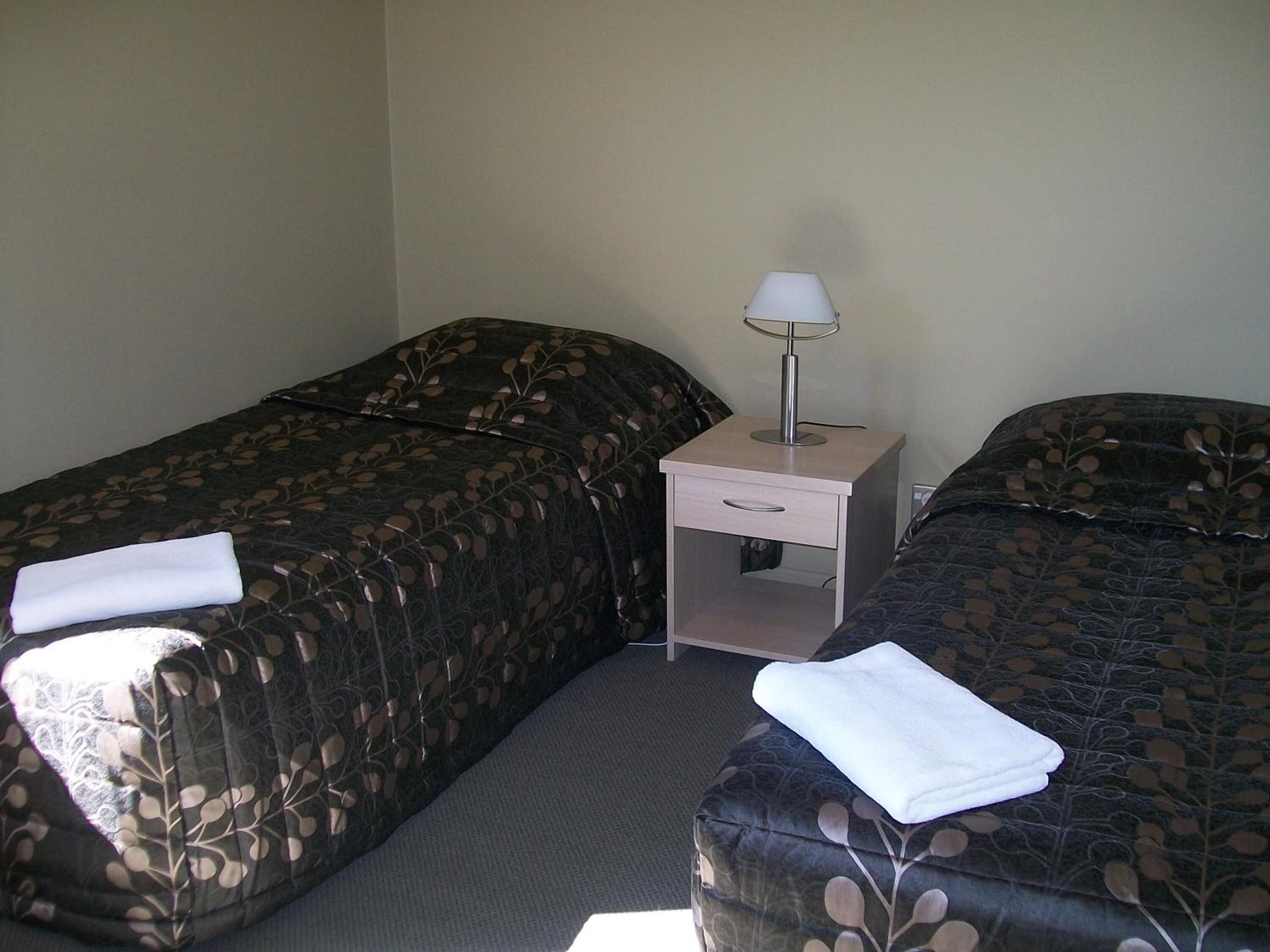 A hotel room with two single beds, a nightstand with a lamp, and a small drawer.