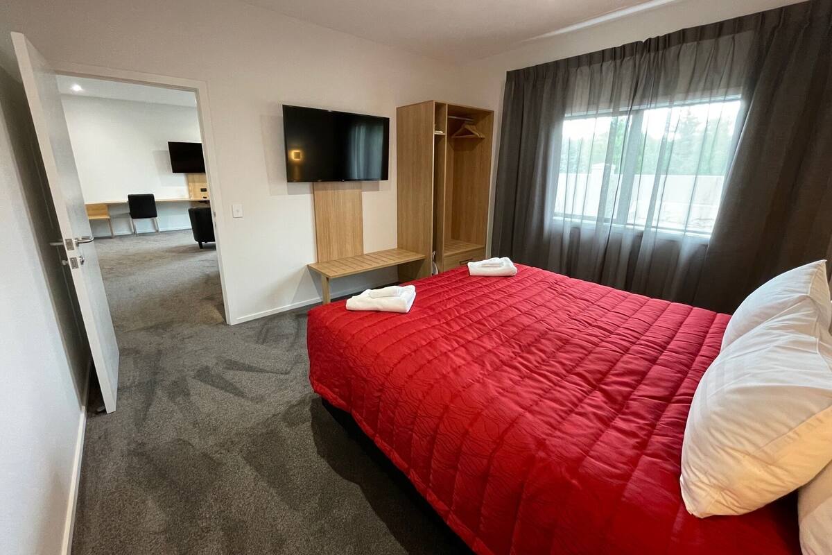 A hotel room with a red bed, a TV mounted on the wall, a wooden wardrobe, and a window with curtains.