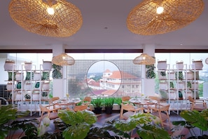 A modern interior with wicker ceiling lights, round tables, chairs, and a view of buildings through a large window. - Wimarion Hotel Semarang (Semarang)