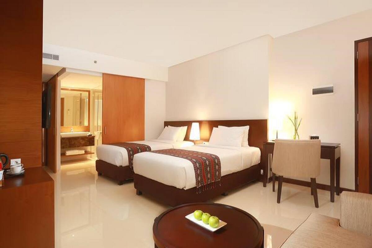 A hotel room with two beds, a desk, a chair, and a round coffee table with apples.